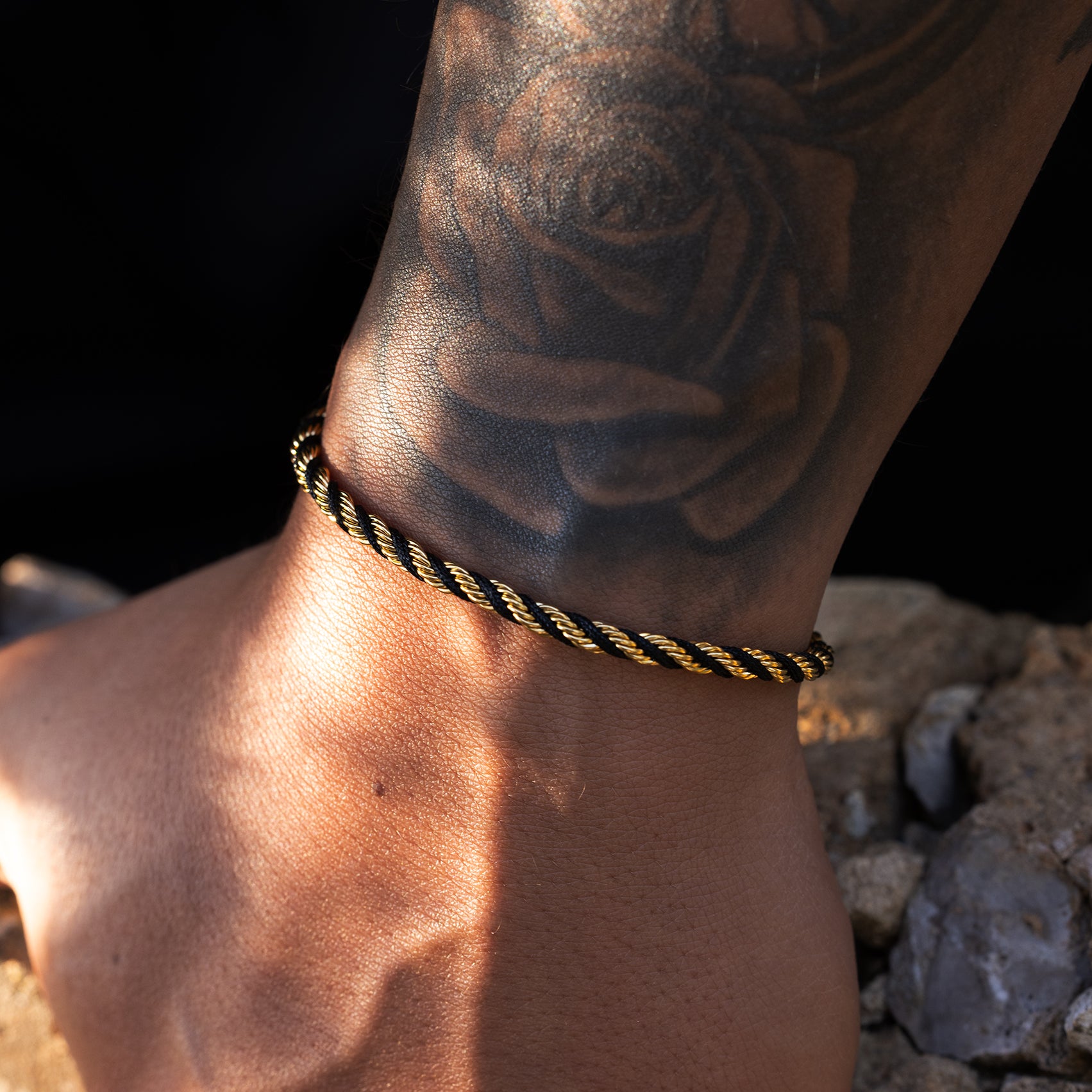 Ankle with a black and gold braided anklet and a tattoo of a rose on a rocky background